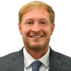 Dr. Daniel Palac smiling, wearing a gray suit jacket, white dress shirt, and patterned tie, against a plain white background.