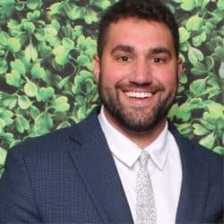 Dr. Jason Cohen smiling in front of a green leafy backdrop, wearing a dark blue suit jacket, white dress shirt, and patterned tie.