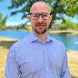 Dr. Adam Taggart smiling outdoors, wearing glasses and a light blue button-down shirt, standing near a river with trees and a bright sky in the background.