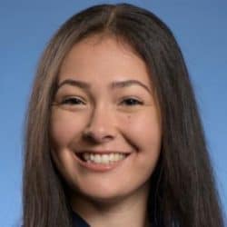 Kelly Ryono pictured with long, straight dark hair smiles warmly at the camera. She has neatly shaped eyebrows, clear skin, and is photographed against a solid light-blue background in a professional-style headshot.