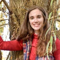 Reilly Ruzella is pictured with long, wavy brown hair smiles while standing among the branches of a tree. She is wearing a red long-sleeve top and a plaid vest, with one arm resting on the tree trunk. Soft sunlight filters through the leaves in the background, creating a warm, natural setting.