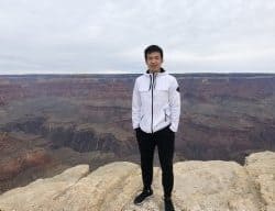 Dr. Yan Luo wearing a white jacket and black pants stands near the edge of a rocky overlook at the Grand Canyon. The sky is overcast, and the layered canyon landscape stretches out behind them.